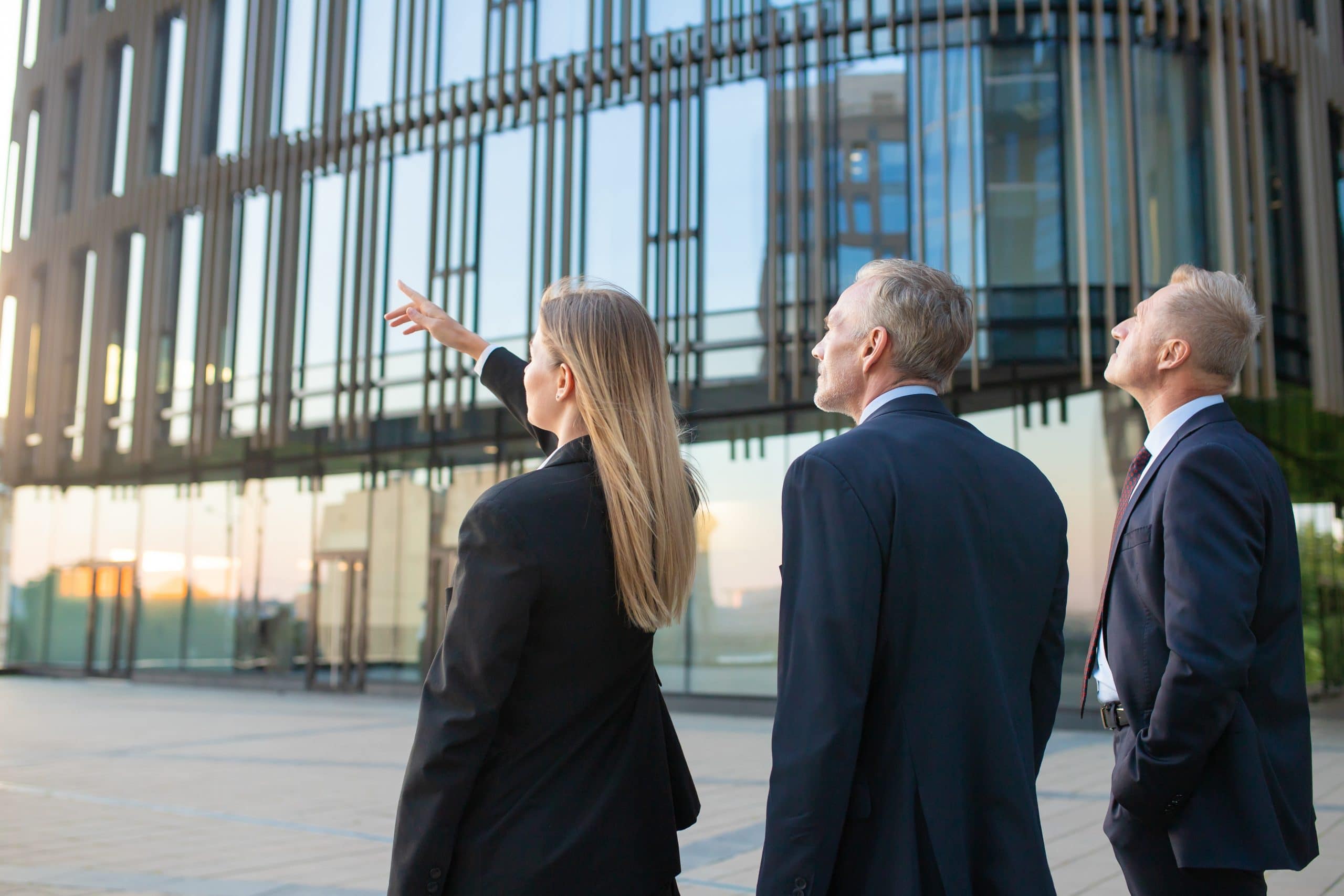 Agent and customers meeting outdoors, discussing real property, pointing at office building. Back view. Commercial real estate concept